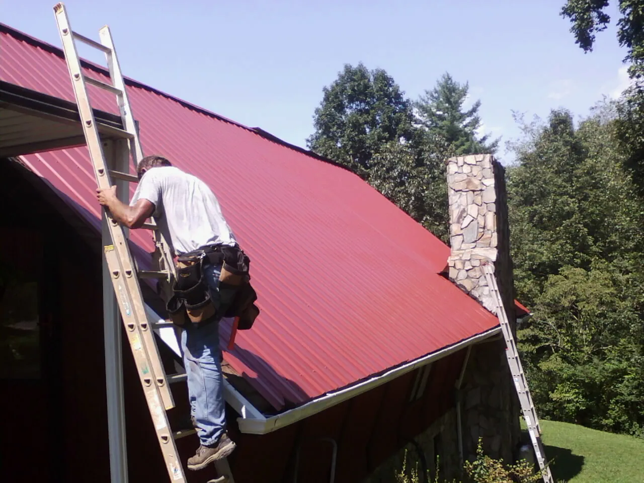 Roofer on a ladder beside a red metal roof and stone chimney — Soffit & Fascia Repair by Roofing Brothers in Campbelltown